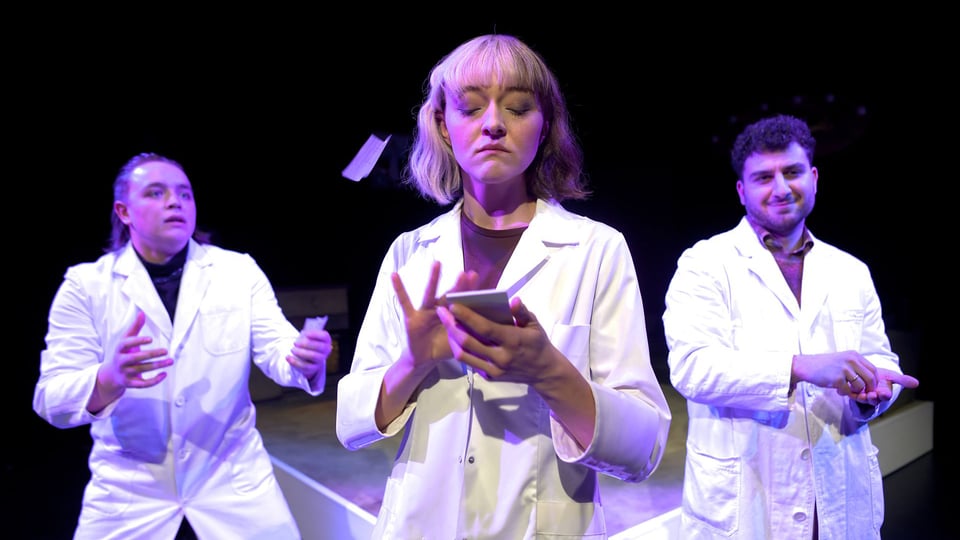 Drei Darsteller*innen in weißen Kitteln, eine Dame mit einem Notizblock