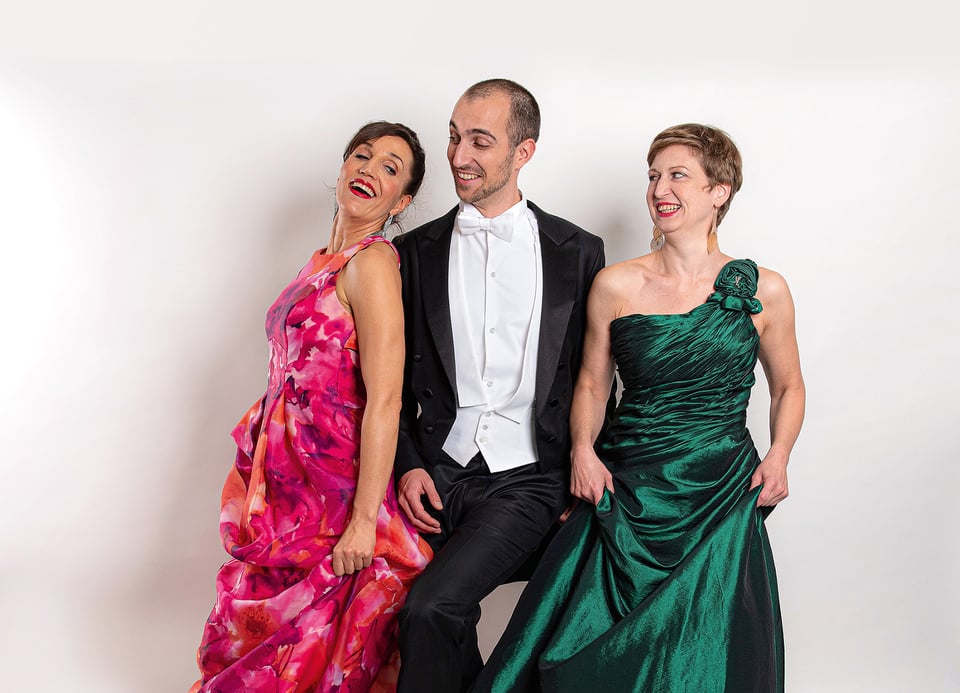 Zwei Damen in rotem und grünem Abendkleid, ein Mann in der Mitte mit schwarzem Anzug und weißem Hemd