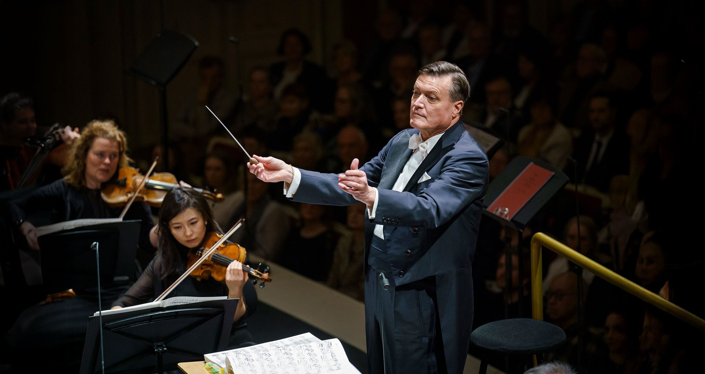 Dirigent in dunkelblauem Anzug neben Orchestermusikern