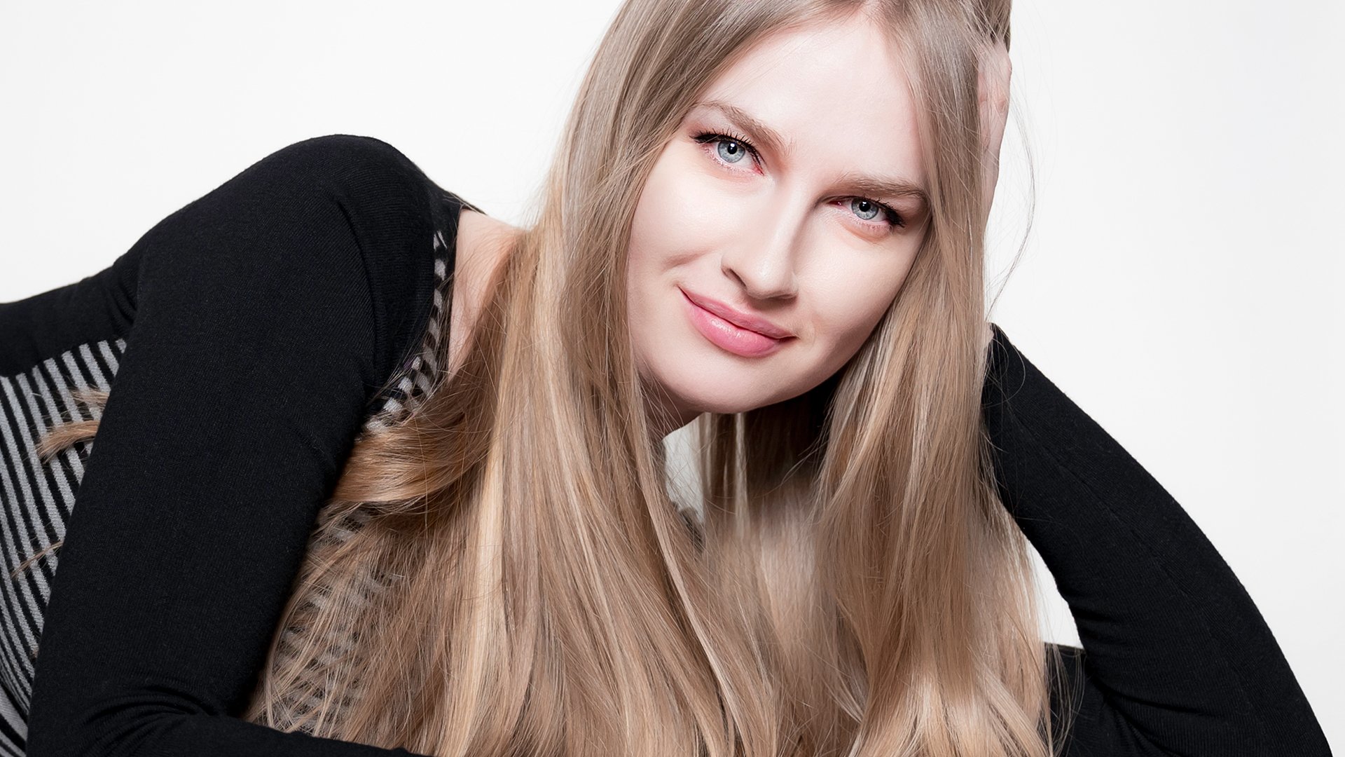 Porträtfoto von Kristina Miller in Nahaufnahme vor hellem Hintergrund, mit langen blonden Haaren und schwarzem Oberteil, den Kopf auf eine Hand gestützt und mit direktem Blick zur Kamera.