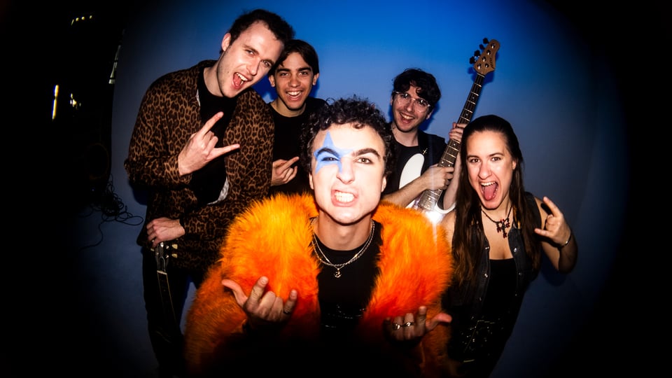 Der Künstler Cosmo mit orangefarbener Jacke und Gesichtsbemalung und mit seiner Band vor dunklem Hintergrund, vier Personen zeigen Handgesten, eine Person hält eine Gitarre hoch 