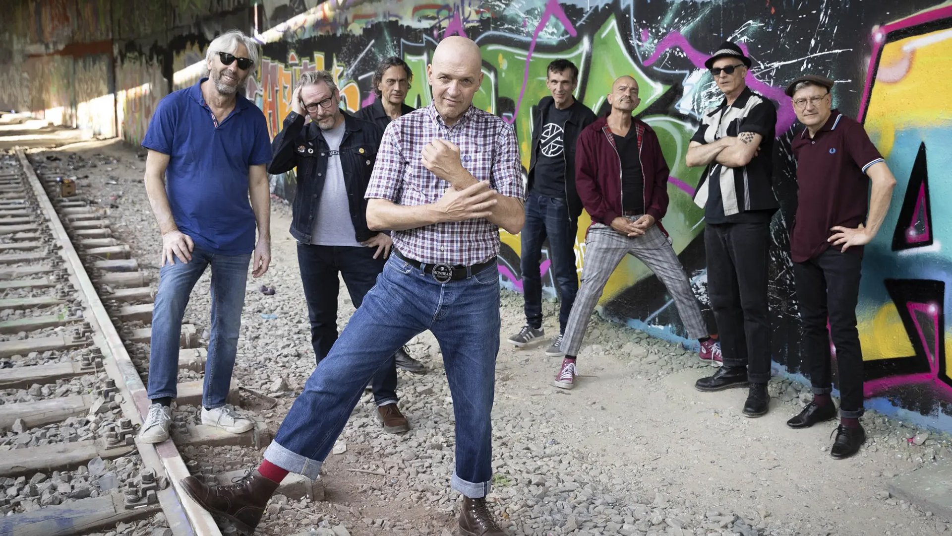 Foto zum 36 Jahre Backstage Jubiläum mit acht Personen in Freizeitkleidung vor einer bunt besprühten Graffitiwand entlang von Bahngleisen, die Gruppe steht verteilt zwischen Schotterbett und Wand, eine Person im karierten Kurzarmhemd posiert im Vordergrund.