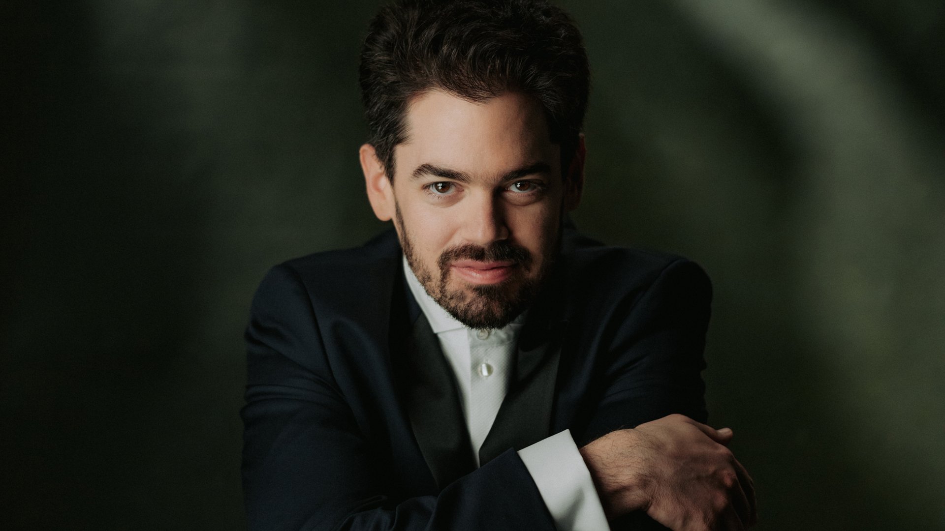 Lahav Shani mit Bart im dunklen Anzug, vor dunkelgrünem Hintergrund.