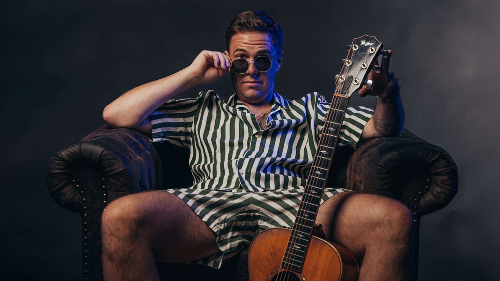 Der Künstler sitzt im gestreifter Kleidung mit Sonnenbrille und Gitarre auf einem antiken Ledersessel