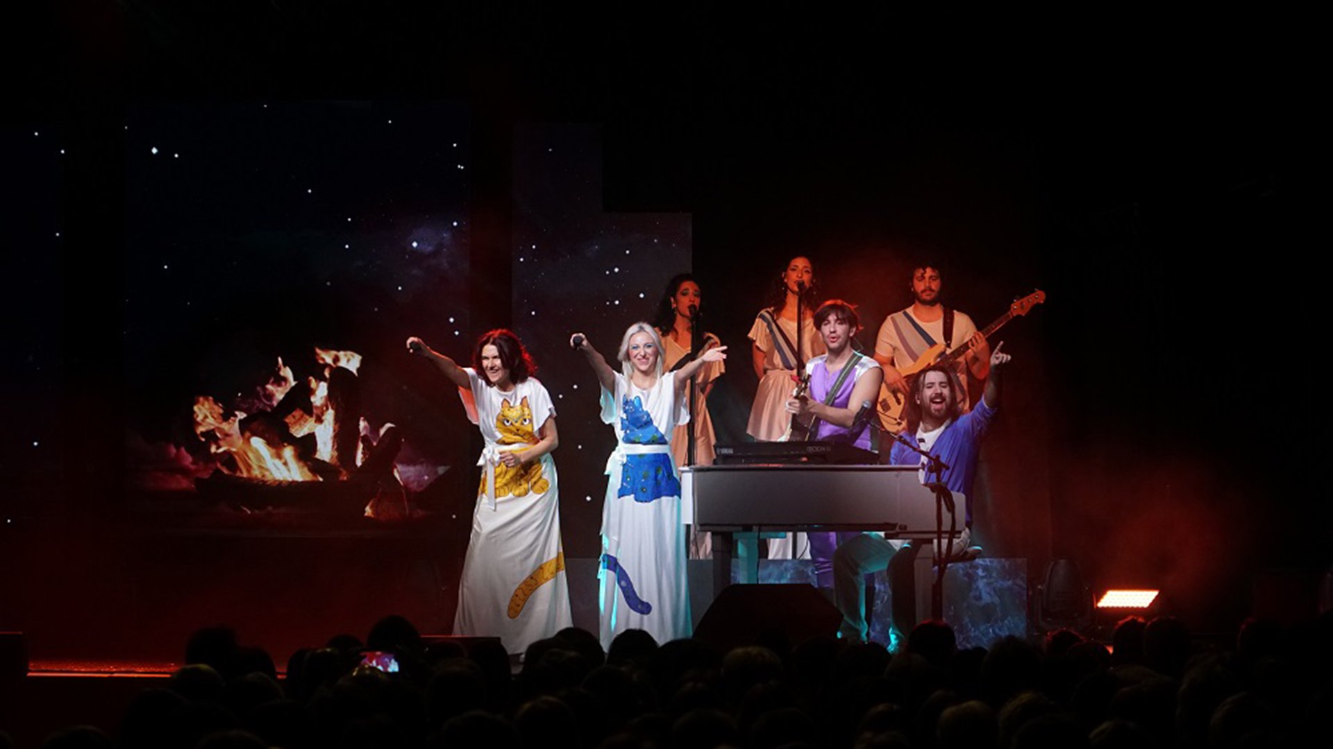 Band auf Bühne im ABBA-Stil mit Sängerinnen in weißen langen Kleidern mit blauem und gelbem Tieraufdruck, Musikern und farbigem Bühnenlicht
