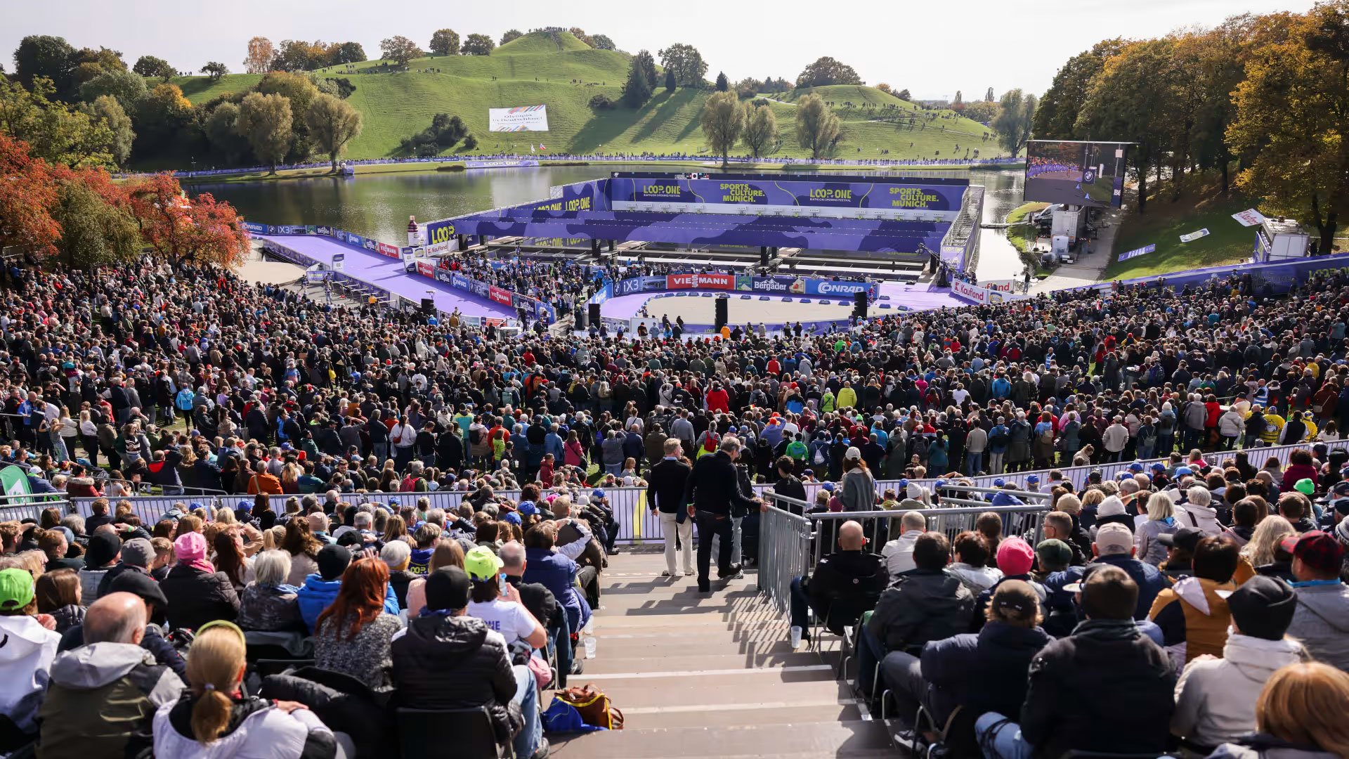 Zuschauerinnen und Zuschauer im Olympiapark München beim LOOP ONE Festival 2026 vor einer Wettkampfarena am See, großes Sportevent unter freiem Himmel mit Tribünen und Bühne.