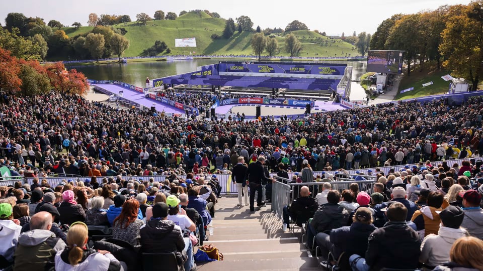 Zuschauerinnen und Zuschauer im Olympiapark München beim LOOP ONE Festival 2026 vor einer Wettkampfarena am See, großes Sportevent unter freiem Himmel mit Tribünen und Bühne.