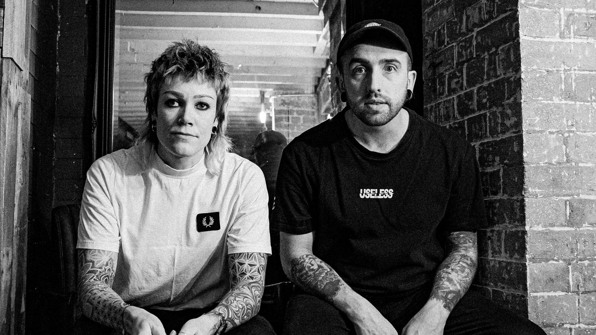 Schwarz-weißes Bandfoto von THE MEFFS. Eine Person mit kurzem Haar trägt ein helles T-Shirt, die andere eine dunkle Kappe und ein schwarzes T-Shirt. Beide sitzen nebeneinander und blicken direkt in die Kamera.