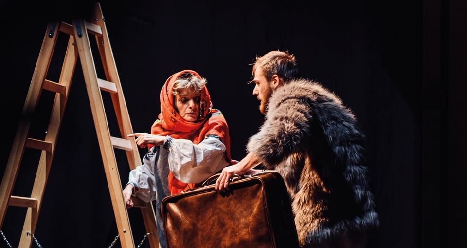 EIne Frau mit orangem Kopftuck und ein Mann mit Pelzjacke und Koffer an einer Leiter stehend