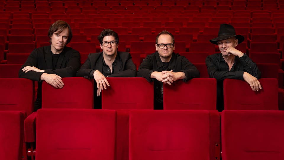Pressefoto von Vokal Total Rock 4 mit vier Musikern in schwarzer Kleidung, die sich über die Rückenlehnen roter Kinosessel lehnen, vor einem leeren Zuschauerraum mit Reihen roter Sitze.
