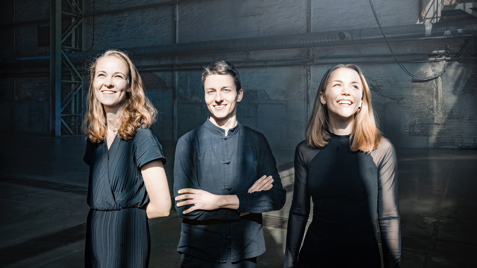 Pressefoto des Amelio Trios mit drei Personen in schwarzer Kleidung, die in einer großen Industriehalle stehen, beleuchtet von einfallendem Tageslicht vor grauen Betonwänden und Stahlträgern.