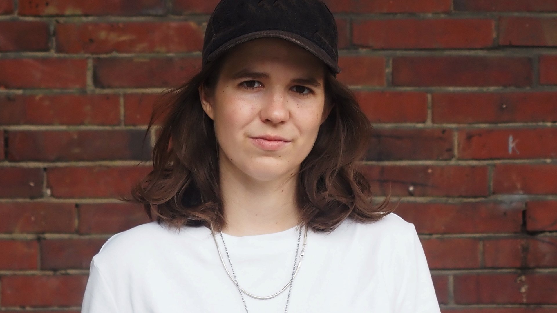 Sophia Merwald mit weißem Shirt und schwarzer Kappe vor einer Ziegelwand