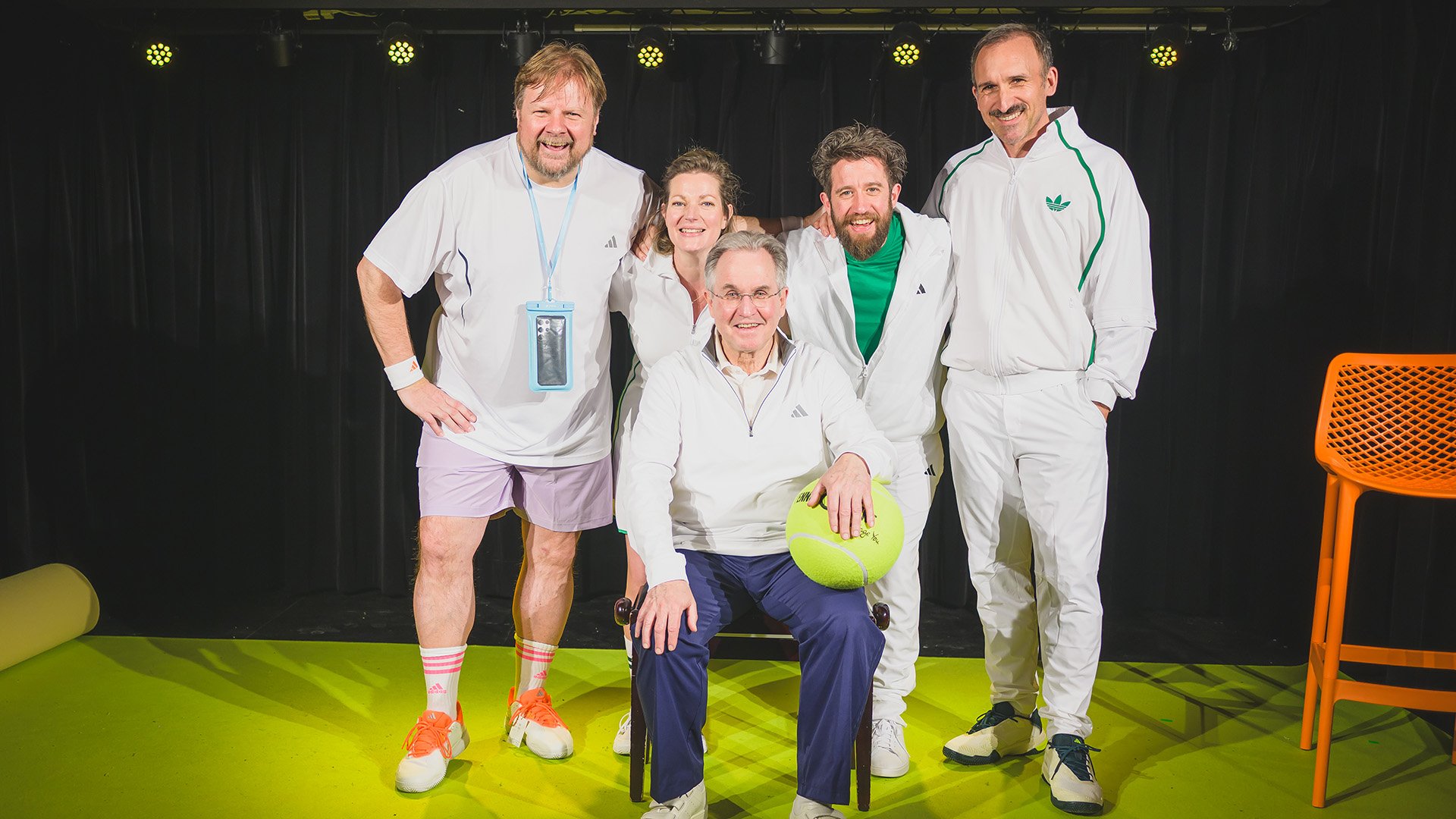 Fünf Personen in weißer Sportbekleidung posieren auf einer Bühnenkulisse, die an einen Tennisplatz erinnert. Eine Person sitzt auf einem Stuhl und hält einen übergroßen Tennisball, während die anderen daneben stehen. Im Hintergrund sind schwarze Vorhänge und Bühnenstrahler zu sehen, rechts steht ein orangefarbener Stuhl.