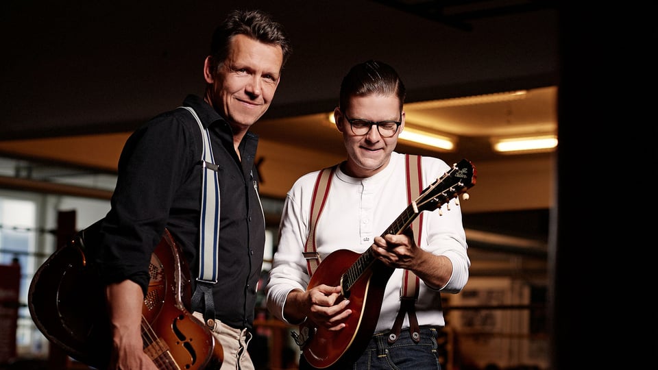 Pressefoto zum Event von Black Patti: Zwei Musiker stehen nebeneinander in einem Innenraum. Eine Person hält eine Resonatorgitarre, die andere spielt eine kleine Saiteninstrument‑Mandoline. Beide tragen Hemden mit Hosenträgern. Die Szene ist warm ausgeleuchtet und vermittelt den Eindruck eines akustischen Blues‑ und Roots‑Konzerts.