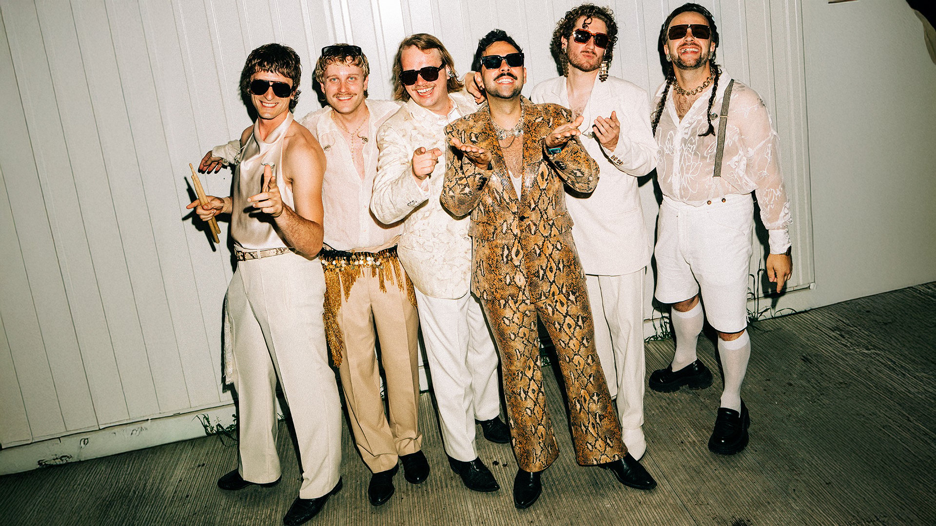 Die sechs Mitglieder von Roy Bianco und die Abbrunzati Boys stehen vor einen weißen Mauer. Sie tragen weiß-beige Bühnenoutfits und Sonnenbrillen.