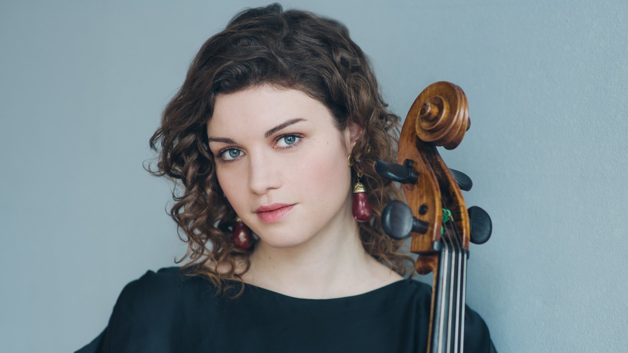 Porträtfoto von Julia Hagen in schwarzer Kleidung, mit Violoncello neben dem Gesicht, vor hellblauem Hintergrund, mit lockigem Haar und rotem Ohrschmuck.