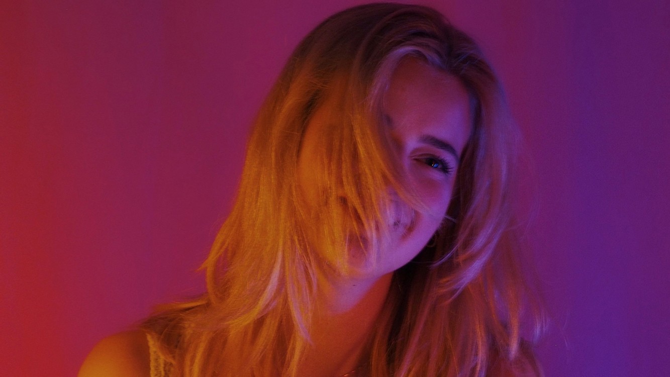 Porträtfoto von Amelie Scheffels mit langen blonden Haaren vor pink violett beleuchtetem Hintergrund, in Nahaufnahme mit seitlich ins Gesicht fallenden Haarsträhnen.