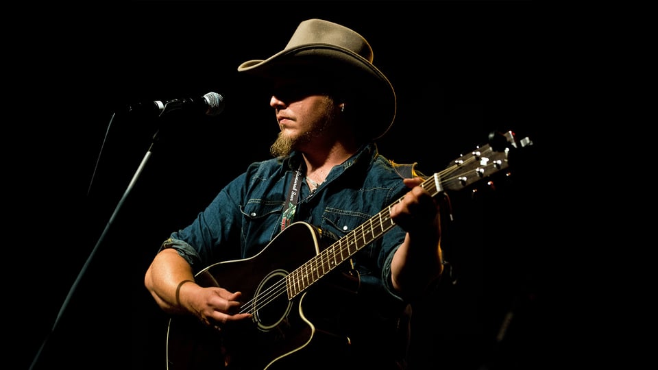 Der Künstler Roman Hofbauer mit Cowboyhut auf spielt Akustikgitarre auf einer dunklen Bühne vor einem Mikrofon und wird von einem einzelnen Scheinwerfer beleuchtet.