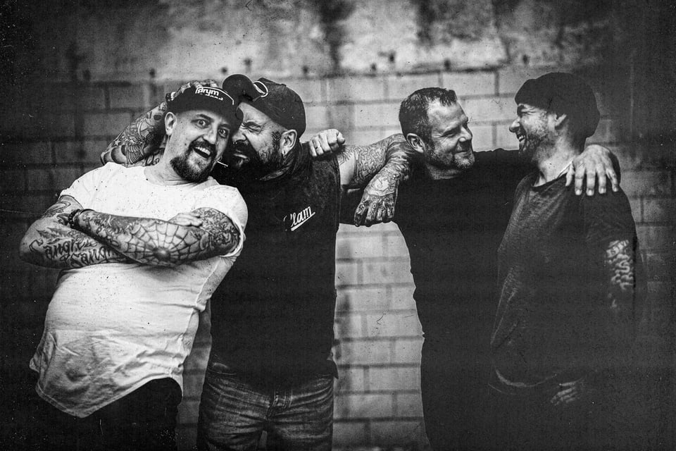 Schwarz-weiss Foto der vier Mitglieder der Band Kärbholz, die nebeneinander stehen und  sich lachend an den Schultern halten, vor rauher Ziegelwand 
