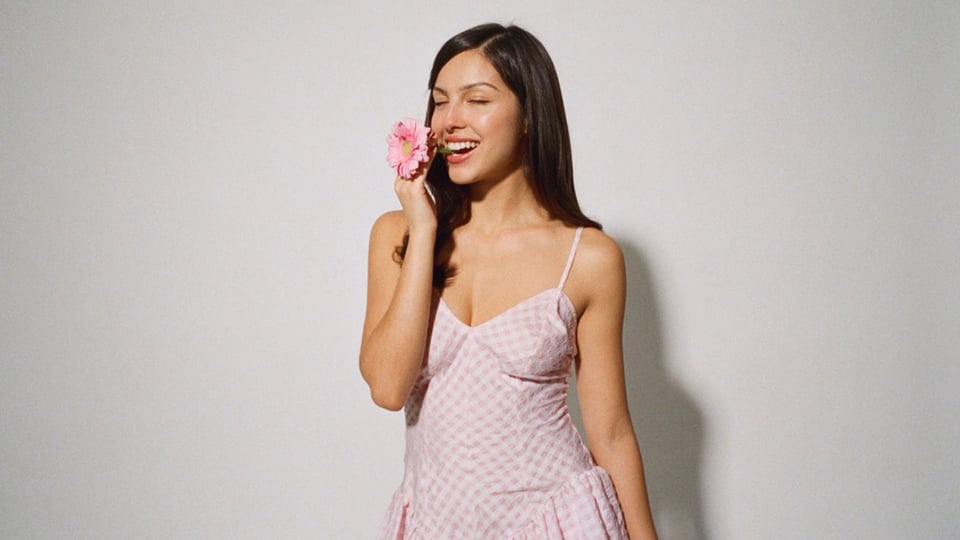 Olivia Rodrigo in rosafarbenem Kleid hält eine Blume an ihr Gesicht vor neutralem Hintergrund, offizielles Pressefoto der Sängerin.
