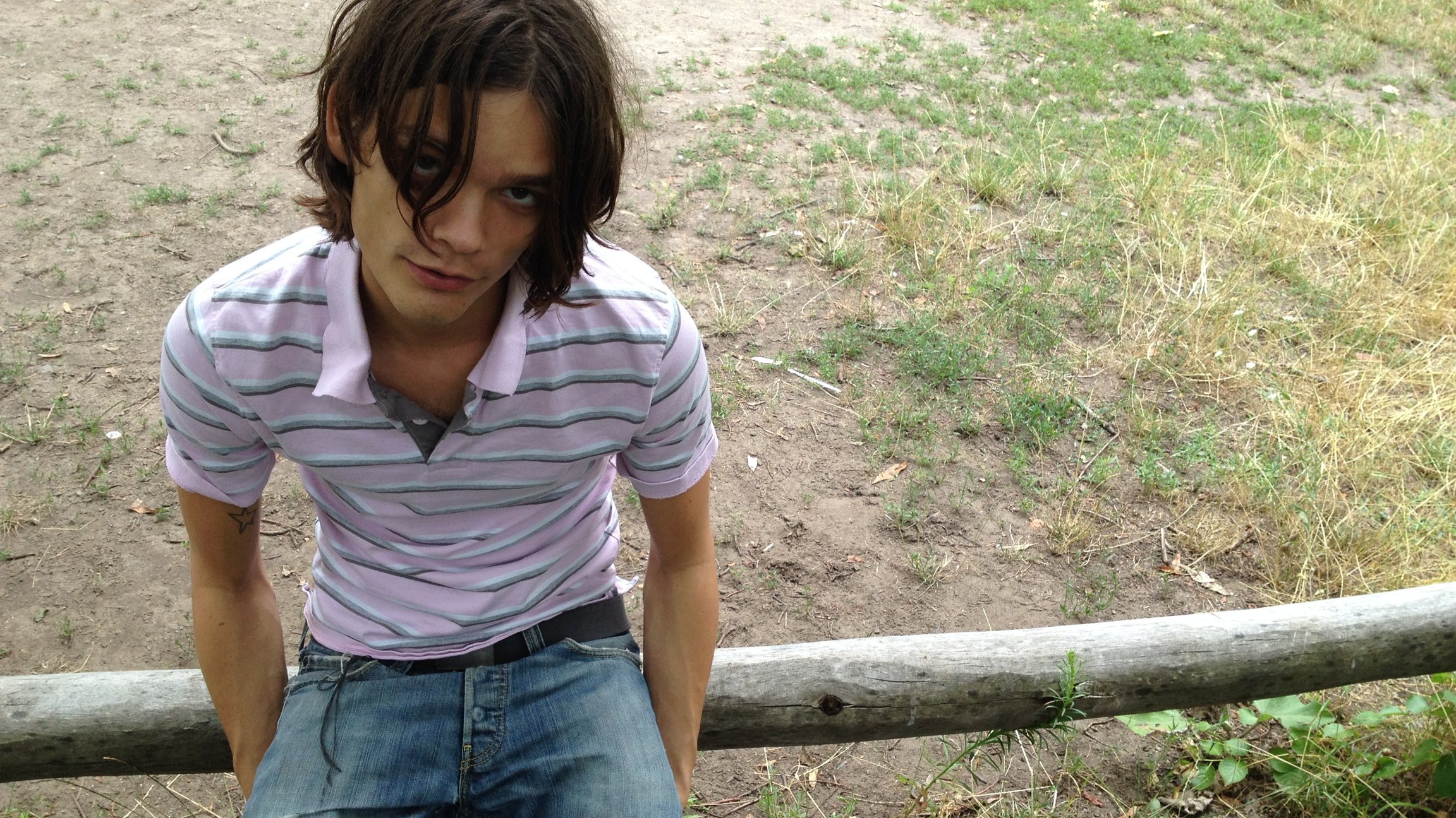 Porträtfoto von Donkey Kid in rosa gestreiftem Poloshirt und Jeans, auf einem Holzgeländer sitzend, mit schulterlangem Haar vor einer trockenen Wiesen- und Erdfläche im Freien.