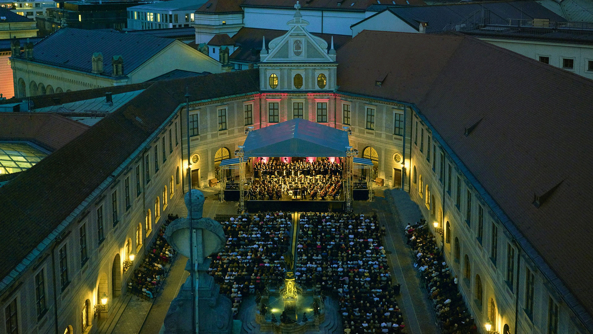 Luftaufnahme eines Open-Air-Konzerts in einem historischen Innenhof bei Abenddämmerung. Vor einer überdachten Bühne mit Orchester und Chor sitzt ein großes Publikum, umgeben von beleuchteten Gebäudefassaden und roter Dachlandschaft.