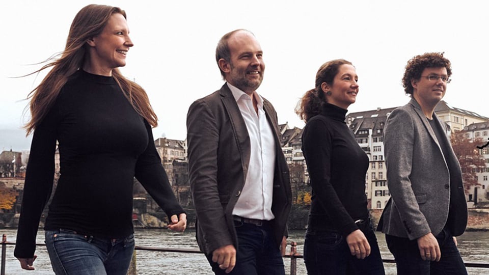 Personengruppe des Amaryllis Quartetts an einer Uferpromenade vor städtischer Kulisse. Vier Ensemblemitglieder gehen nebeneinander am Fluss entlang, im Hintergrund sind Gebäude am gegenüberliegenden Ufer zu sehen.