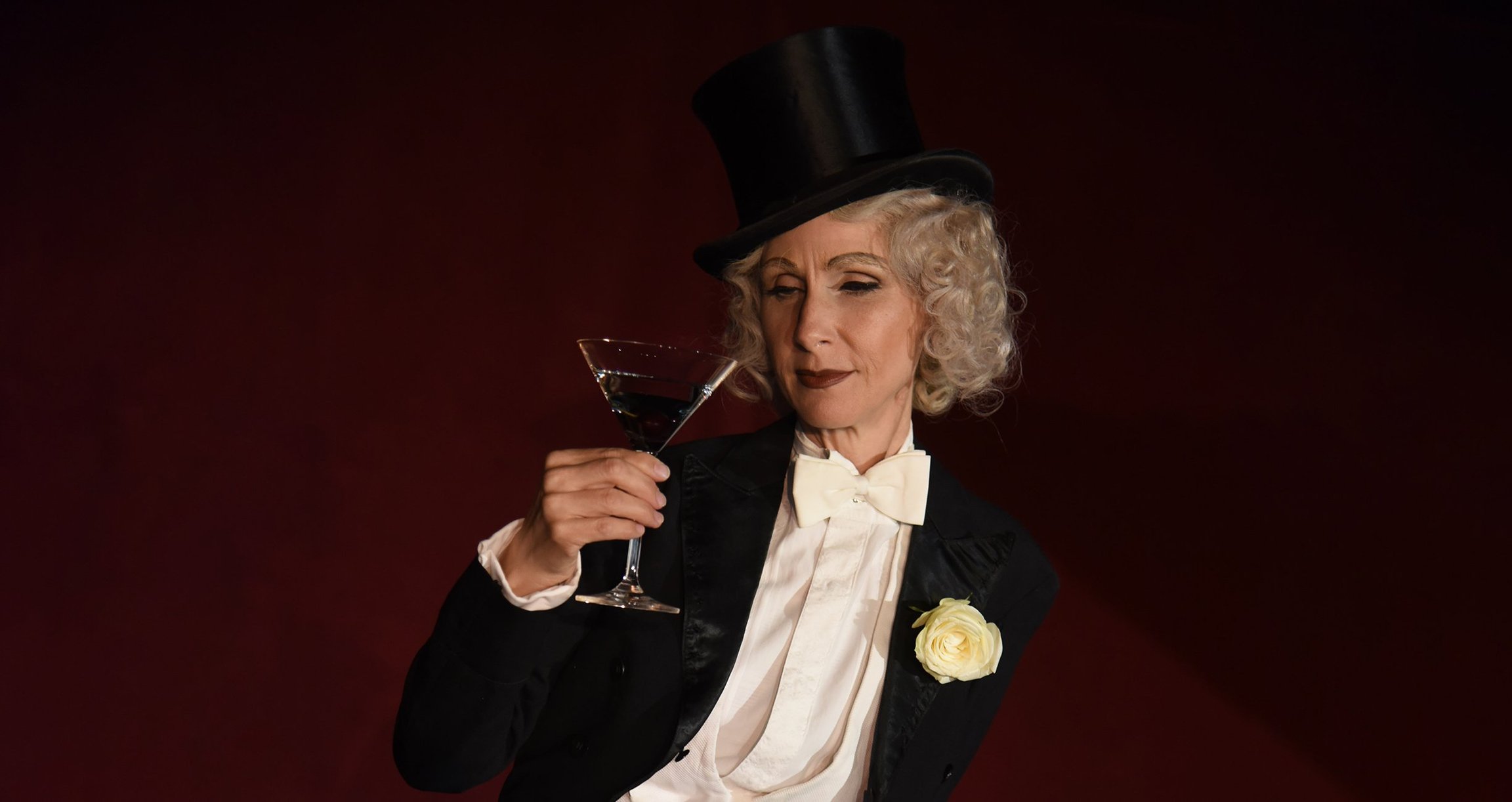 Foto der Künstlerin mit einem Zylinderhut und einem Glas in der Hand, vor einem weinroten Hintergrund