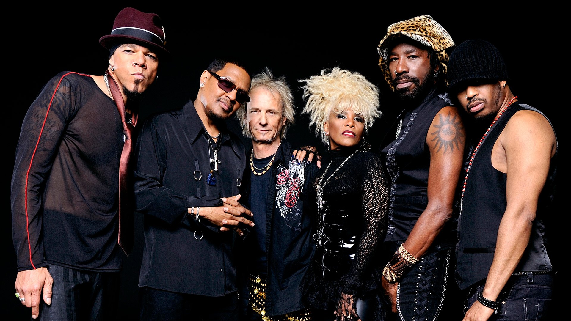 Bandfoto von Mother’s Finest mit sechs Mitgliedern in dunklen Bühnenoutfits vor schwarzem Hintergrund.