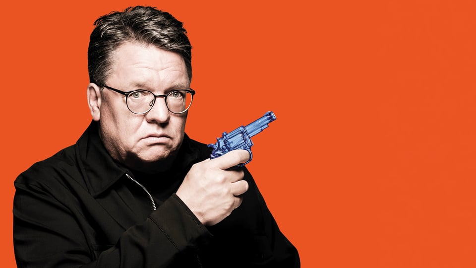 Porträt von Helmut Schleich mit Brille vor orangefarbenem Hintergrund. Die Person hält einen blauen Spielzeugrevolver in der Hand und blickt direkt in die Kamera.