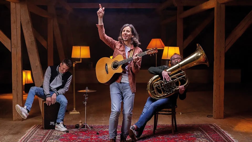 Bandfoto von Jannemacher mit drei Musikern in einer Scheune oder Holzhalle auf orientalischem Teppich, eine Person mit Akustikgitarre im Zentrum, links eine Person auf einer Cajón und rechts eine Person mit Tuba, umgeben von warm beleuchteten Stehlampen und Holzbalken.