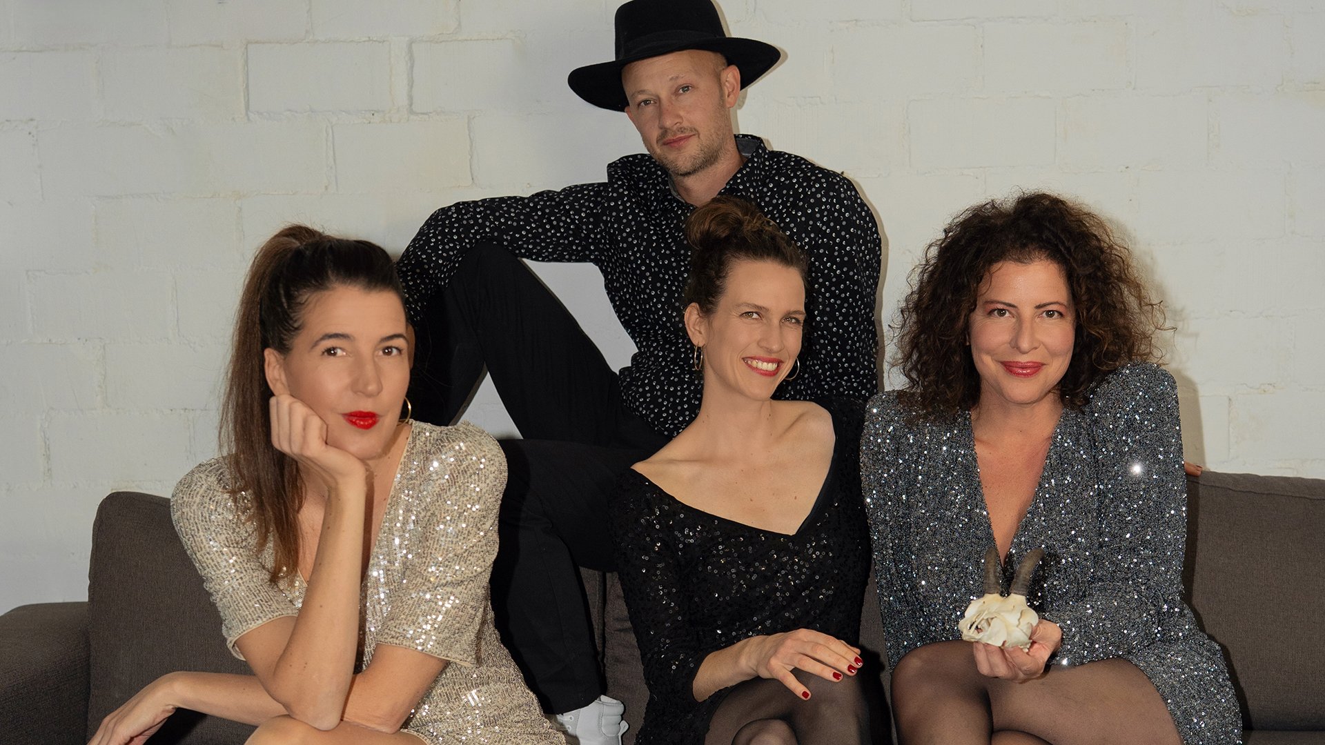 Drei Damen festlich gekleidet und ein Herr mit schwarzem Hut auf einem braunen Sofa vor hellem Hintergund