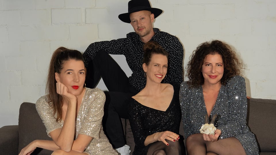 Drei Damen festlich gekleidet und ein Herr mit schwarzem Hut auf einem braunen Sofa vor hellem Hintergund