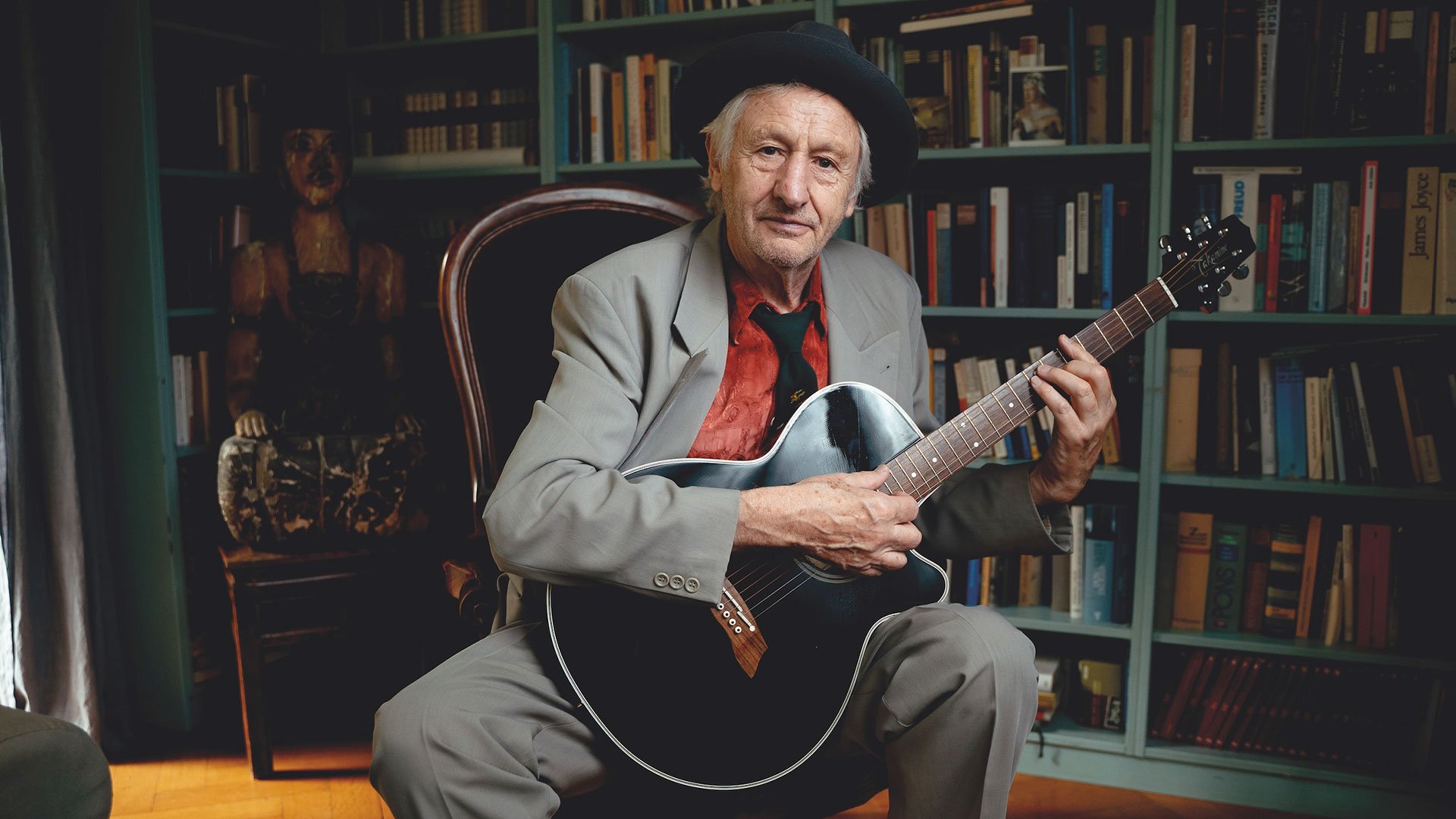 Foto für die Veranstaltung „Georg Ringsgwandl“ mit dem Künstler, der mit Hut auf einem Stuhl sitzt und eine Gitarre in der Hand hält.