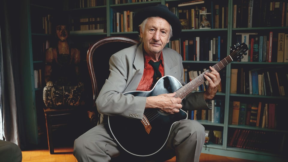 Foto für die Veranstaltung „Georg Ringsgwandl“ mit dem Künstler, der mit Hut auf einem Stuhl sitzt und eine Gitarre in der Hand hält.