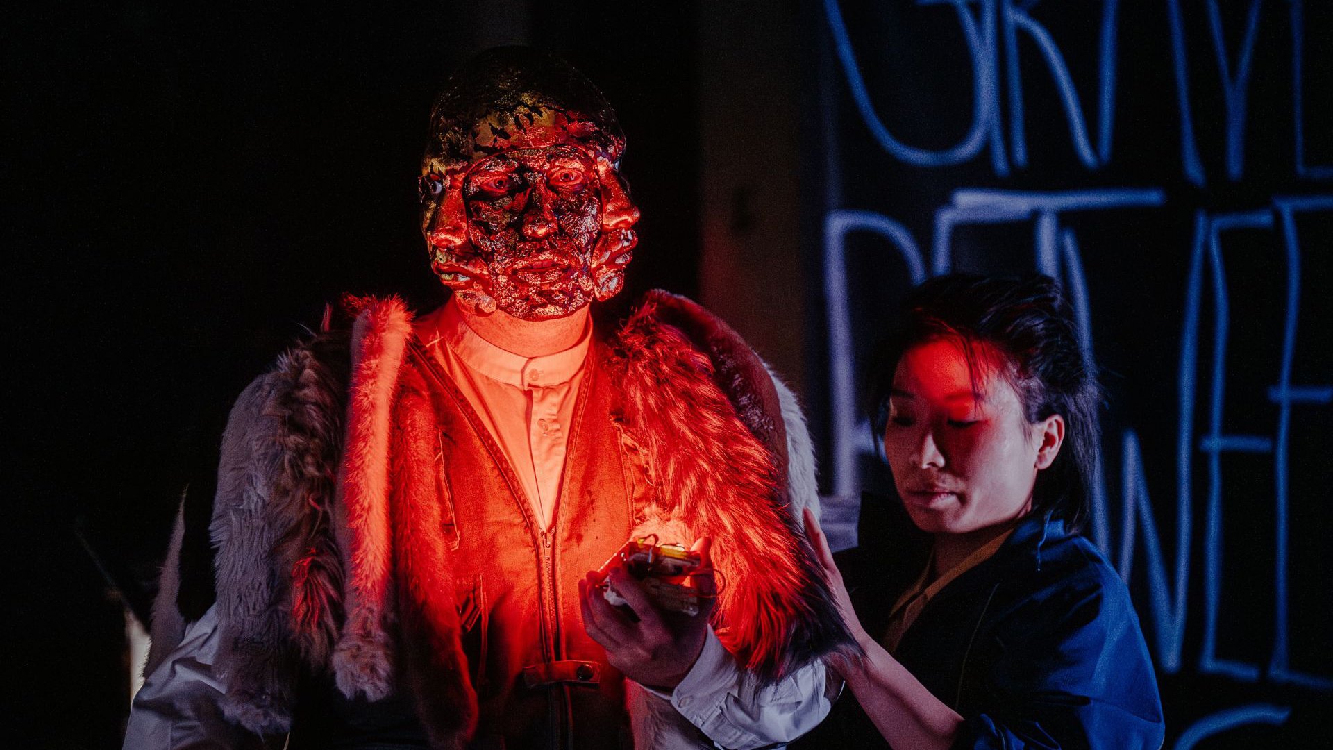 Seine Szene aus Antigone mit zwei Darstellern, eine mit einer Maske mit drei Gesichtern, rot erleuchtet und eine weitere dunkelhaarige Person, im Hintergrund große Buchstaben