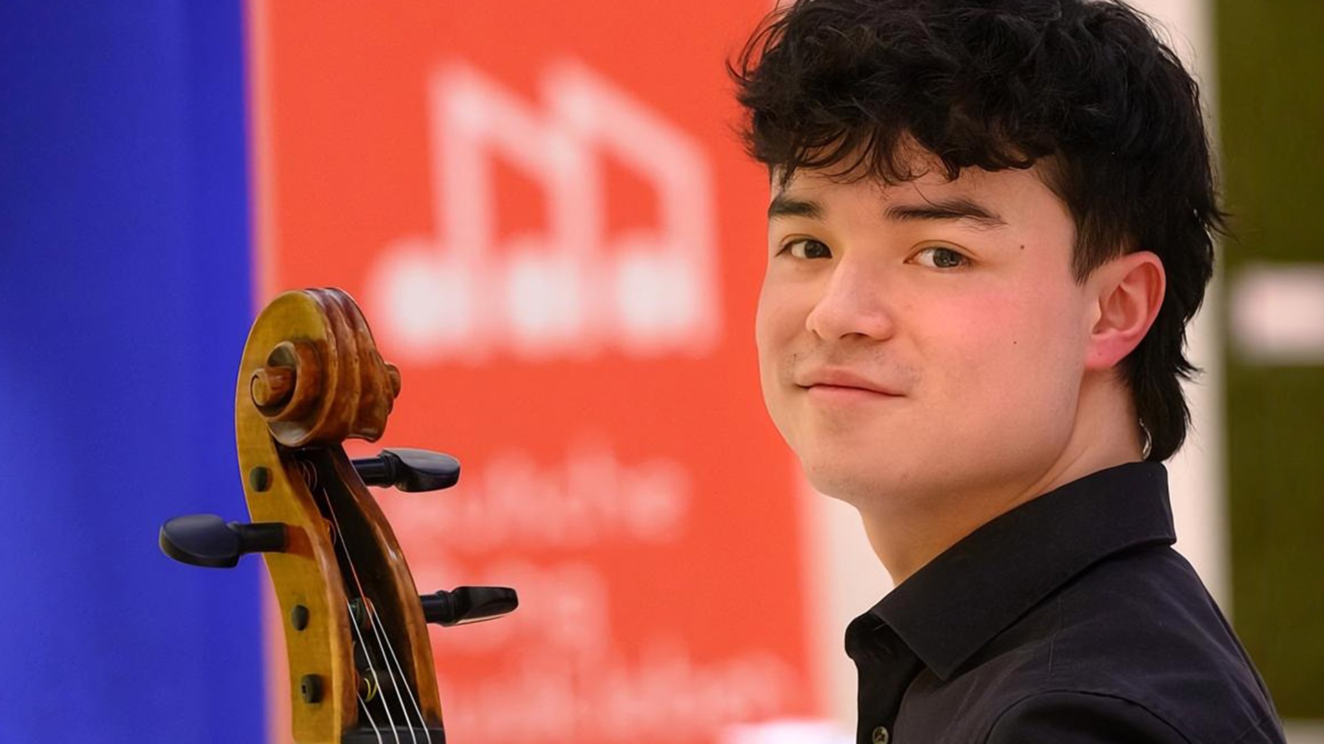 Der Künstler Akito Marschik mit dem Hals eines Streichinstrumentes vor rot-blau-grünem Hintergrund blickt lächelnd in die Kamera