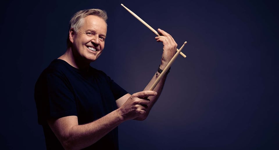 Ein Foto des Künstlers mit Drumsticks vor einem blauen Hintergrund.