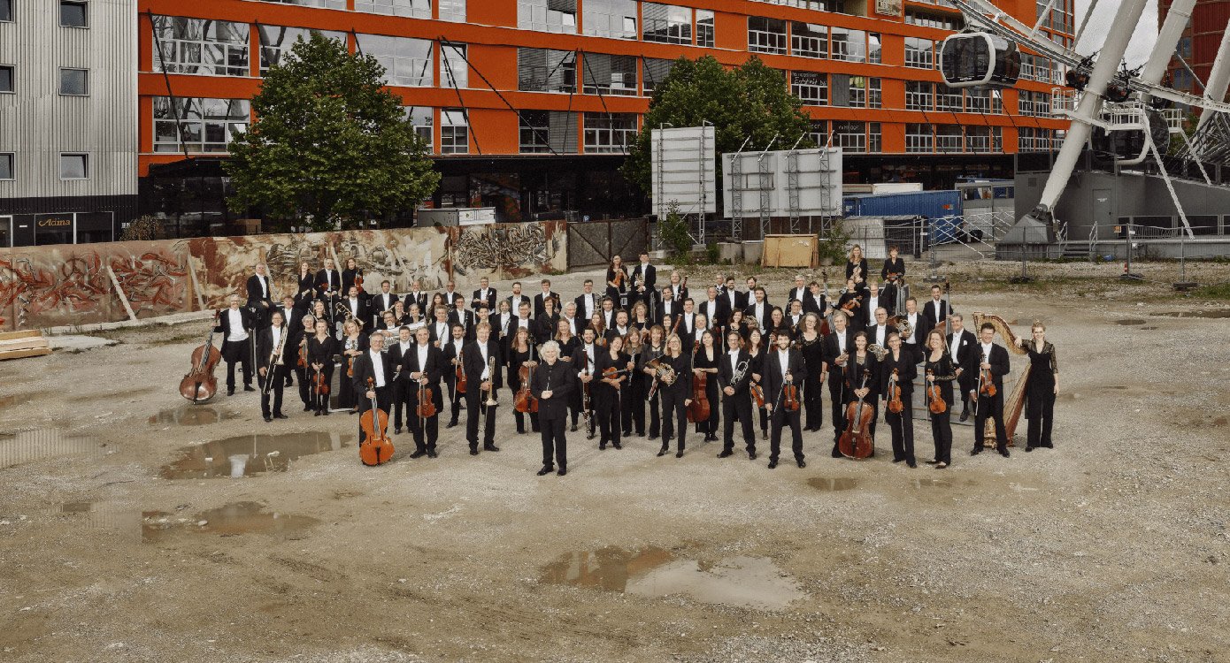 Das Symphonieorchester in dunkler Kleidung mit Instrumenten in Hof des Werksviertels stehend, im Hintergrund Gebäude