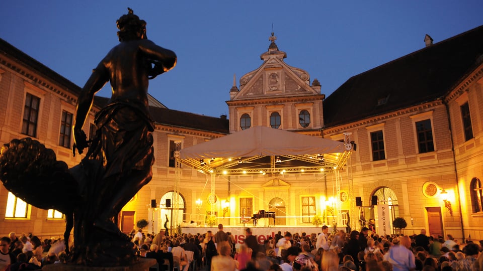 Abendliche Konzertszene im Brunnenhof der Münchner Residenz mit beleuchteter Bühne und großem sitzendem Publikum; im Vordergrund eine Statue, im Hintergrund historisches Gebäude im warmen Licht.