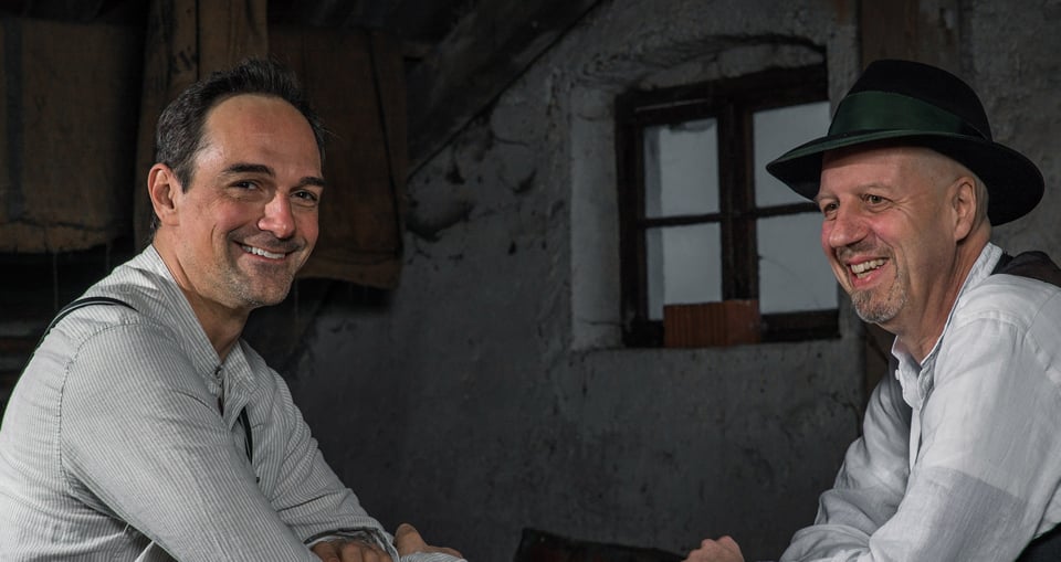 Zwei Männer in Trachtenkleidung, einer mit Hut, vor Hüttenfenster sitzend