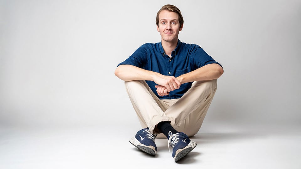 Foto zum Event ‚Jonas Greiner – Auf Augenhöhe‘: Der Künstler sitzt auf dem Boden vor grauem Hintergrund.