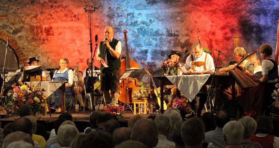 Ein Foto von einem Konzert mit Publikum, auf der Bühne einige Personen mit Instrumenten und in bayerischer Tracht