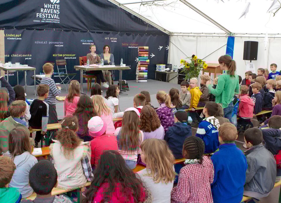 Viele Kinder sitzen auf mehreren Bänken vor einer Bühne in einem Zelt. Auf der Bühne steht ein Tisch an dem zwei Frauen sitzen. Eine Frau spricht durch ein Mikrofon.