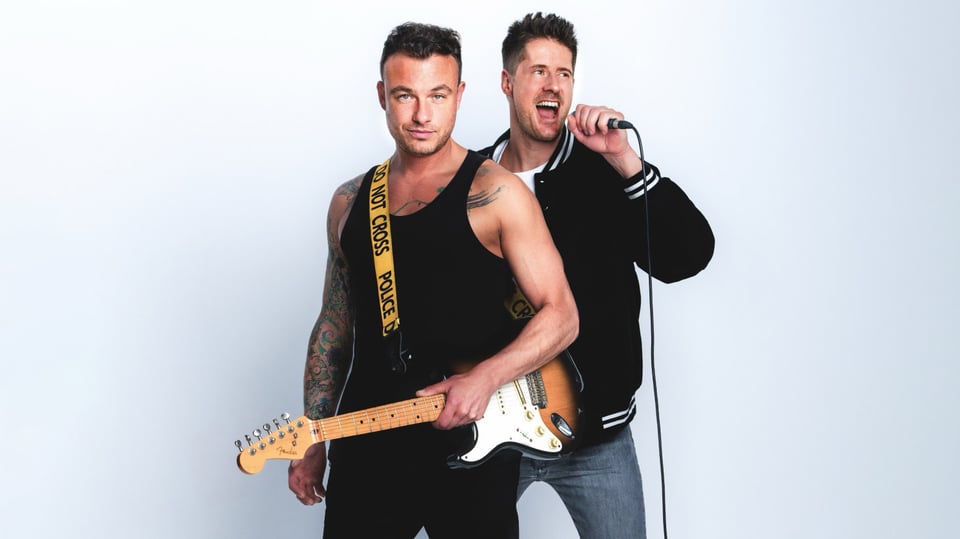Zwei Männer vor einem hellen Hintergrund, von denen einer eine E-Gitarre hält und der andere in ein Mikro singt