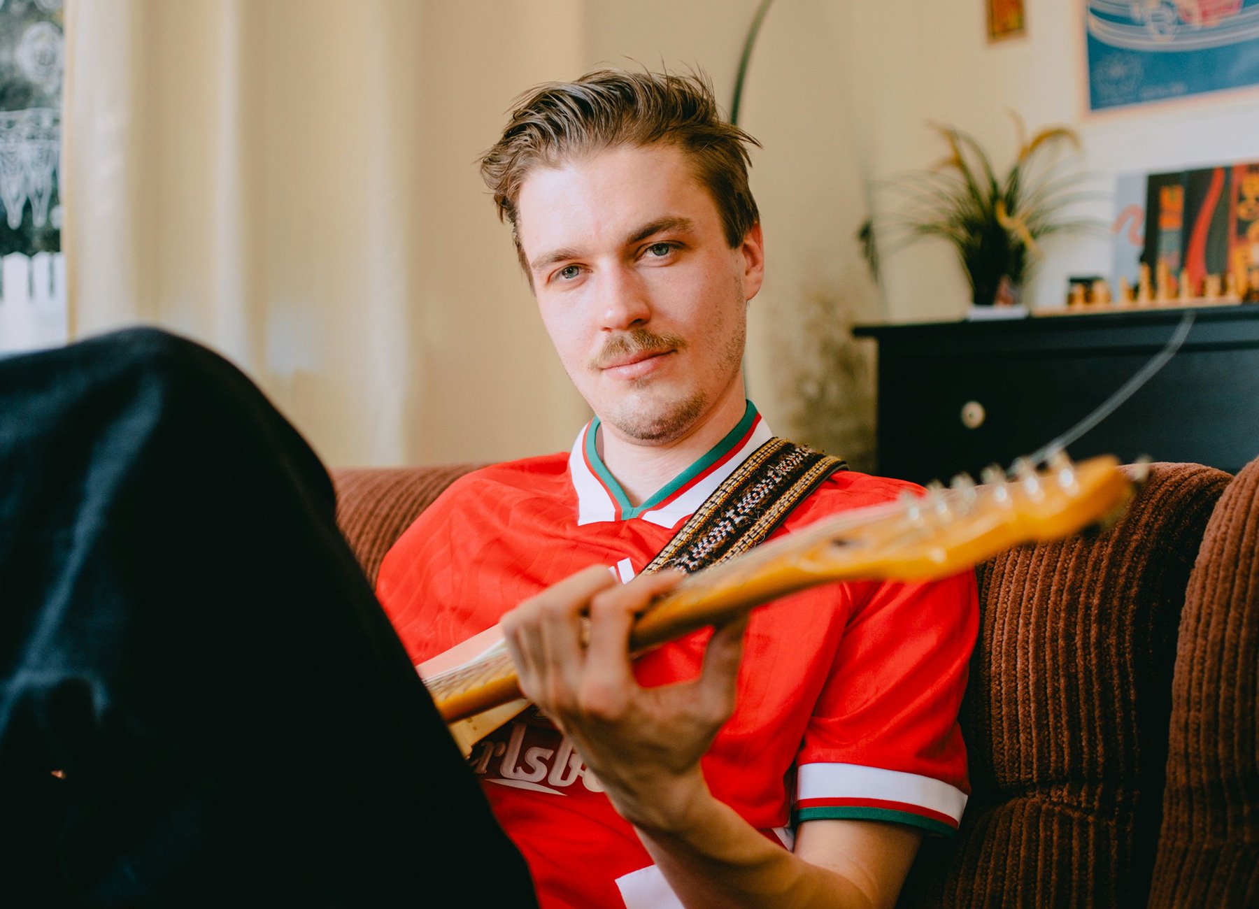 Mann in orangem Shirt mit Gitarre auf Sofa sitzend