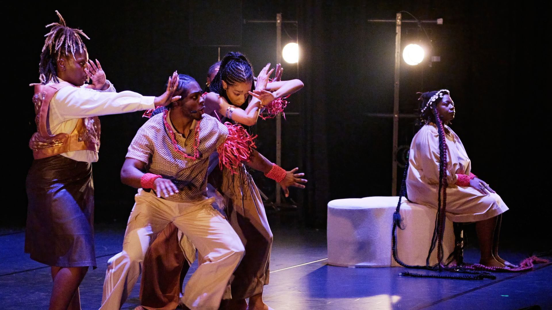 Mehrere Personen in choreografierter Bewegung aus der Veranstaltung OPERA OF HOPE auf einer Bühne, mit ausdrucksstarken Gesten, Kostümen und heller Bühnenbeleuchtung; eine Person sitzt seitlich auf einem Podest.