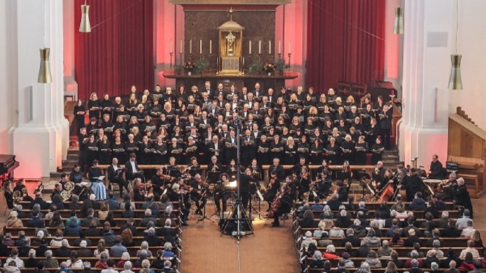Bühnenfoto des Joseph-Chors München mit großem Chor und Orchester vor Publikum in einer Kirche, vor rotem Vorhang und Altarbereich mit Kerzen, während der Dirigent zentral vor dem Ensemble steht.