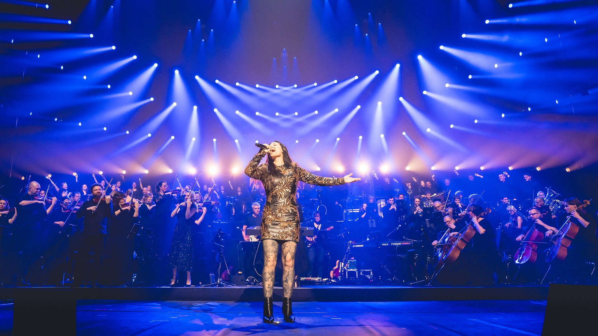 Musikerin singt auf einer großen Bühne vor einem Orchester, trägt ein glitzerndes Kleid und hält ein Mikrofon, umgeben von vielen blauen Scheinwerfern.