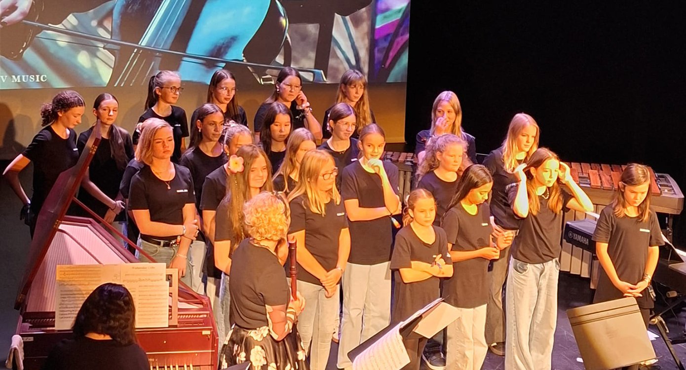 Foto eines Mädchenchors mit einer Frau am Klavier.
